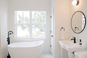A bathroom with a freestanding bathtub, a sink, a mirror, and a window with a view of trees. - Modern home with lookout in the heart of downtown (Wilmington)