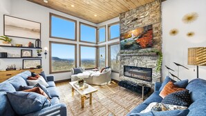 Amplia sala de estar con chimenea de piedra, ventanas grandes y vistas al paisaje.