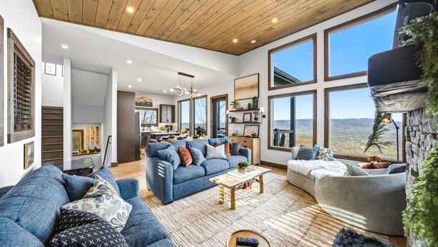 Un salón espacioso con un sofá azul grande, un sofá gris más pequeño y una mesa de centro de madera. Amplias ventanas ofrecen vistas al exterior.