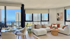 A modern living room with a large window, a sofa, a coffee table, and a dining area. -  Coastal Luxe Apartment with Ocean & River Views (Surfers Paradise)