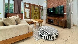 A modern living room with a brick wall, a large flat-screen TV, a sofa, a coffee table, and a patterned ottoman. - Villa White Orchid - Enclosed living area - Seminyak center - Quiet Private Pool (Seminyak)