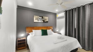 Une chambre d’hôtel moderne avec une tête de lit en bois, un grand lit avec une literie blanche, un coussin vert en guise d’élément décoratif, un ventilateur de plafond et une œuvre d’art représentant un paysage urbain accrochée au mur.