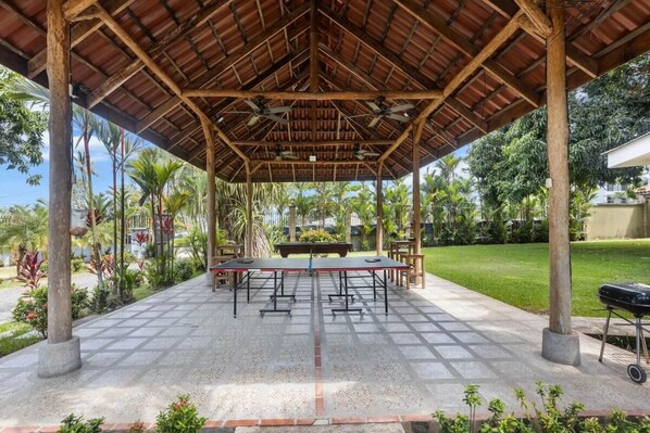 An outdoor covered patio area with a long table and chairs, surrounded by lush greenery and a clear sky.