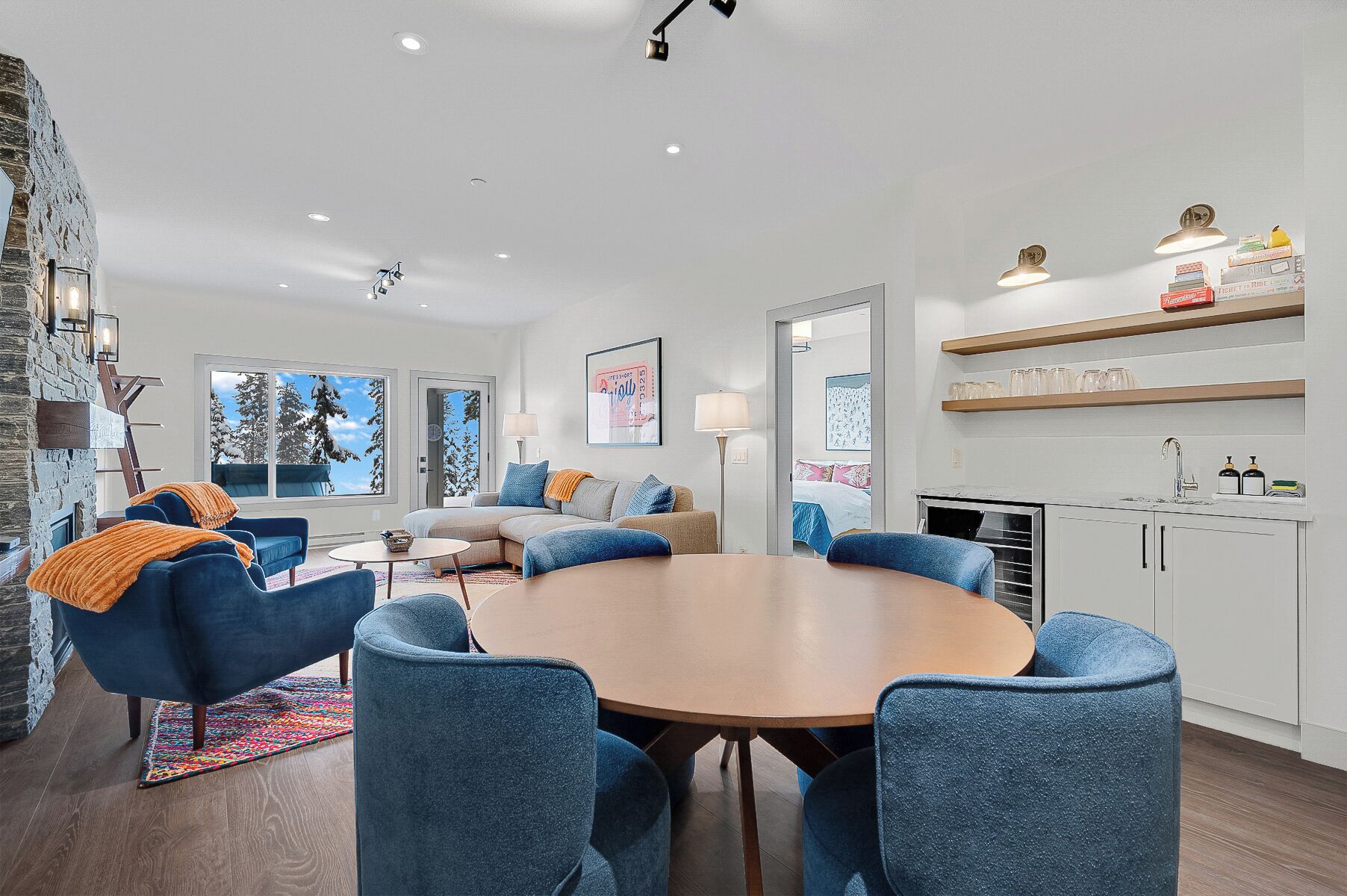 Een moderne woonkamer met een ronde houten tafel, blauwe fauteuils en een bank. Er is een keukenhoek met witte kasten en een aanrecht.