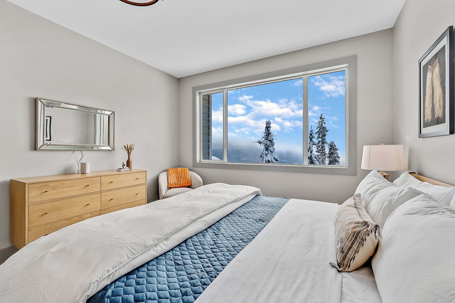 Ein Schlafzimmer mit großem Fenster, einem Holzkonsolen-Schrank, einem Nachttisch und einem Bett mit weißer und blauer Bettwäsche.