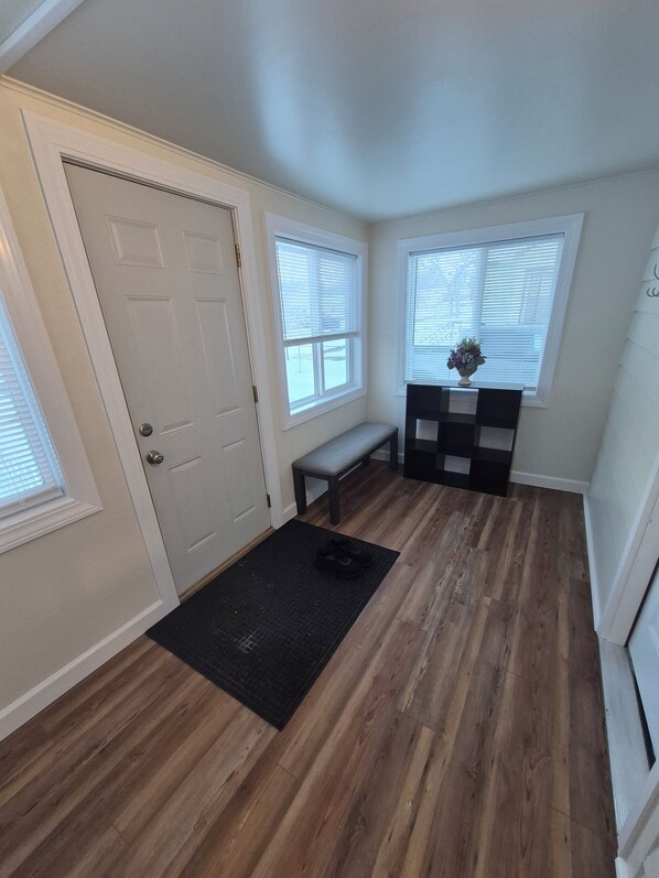 A room with a wooden floor, a bench, a door, and two windows with blinds.