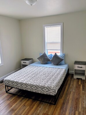 A bedroom with a bed, two nightstands, a window with blinds, and wooden flooring.