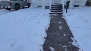A snow-covered pathway leading to a single-story house with a garage, a mailbox, and a parked truck.