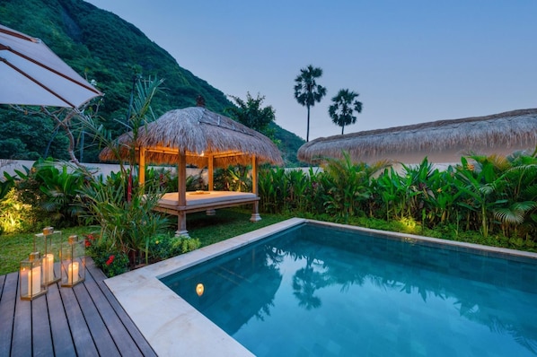 A pool area with a thatched gazebo, lush greenery, and a mountain backdrop.