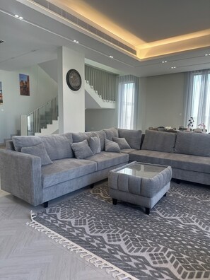 A modern living room with a grey sectional sofa, a matching ottoman, and a patterned rug. A staircase with a glass railing is visible in the background.