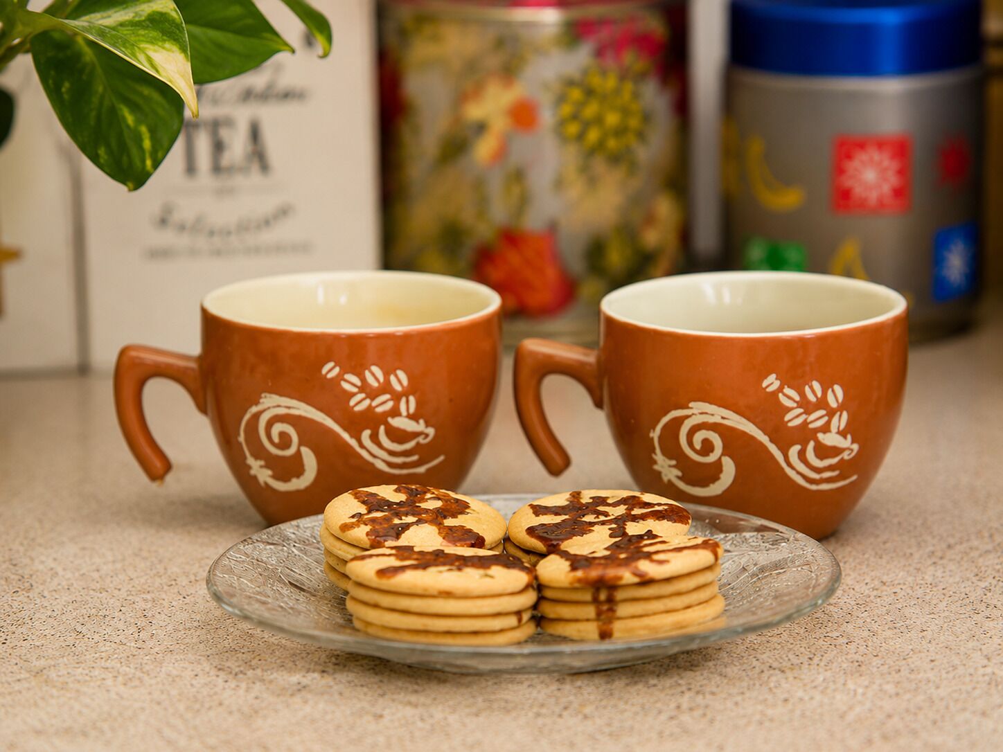 Dos tazas de cerámica marrón con diseños blancos, un plato con galletas y un recipiente en el fondo.