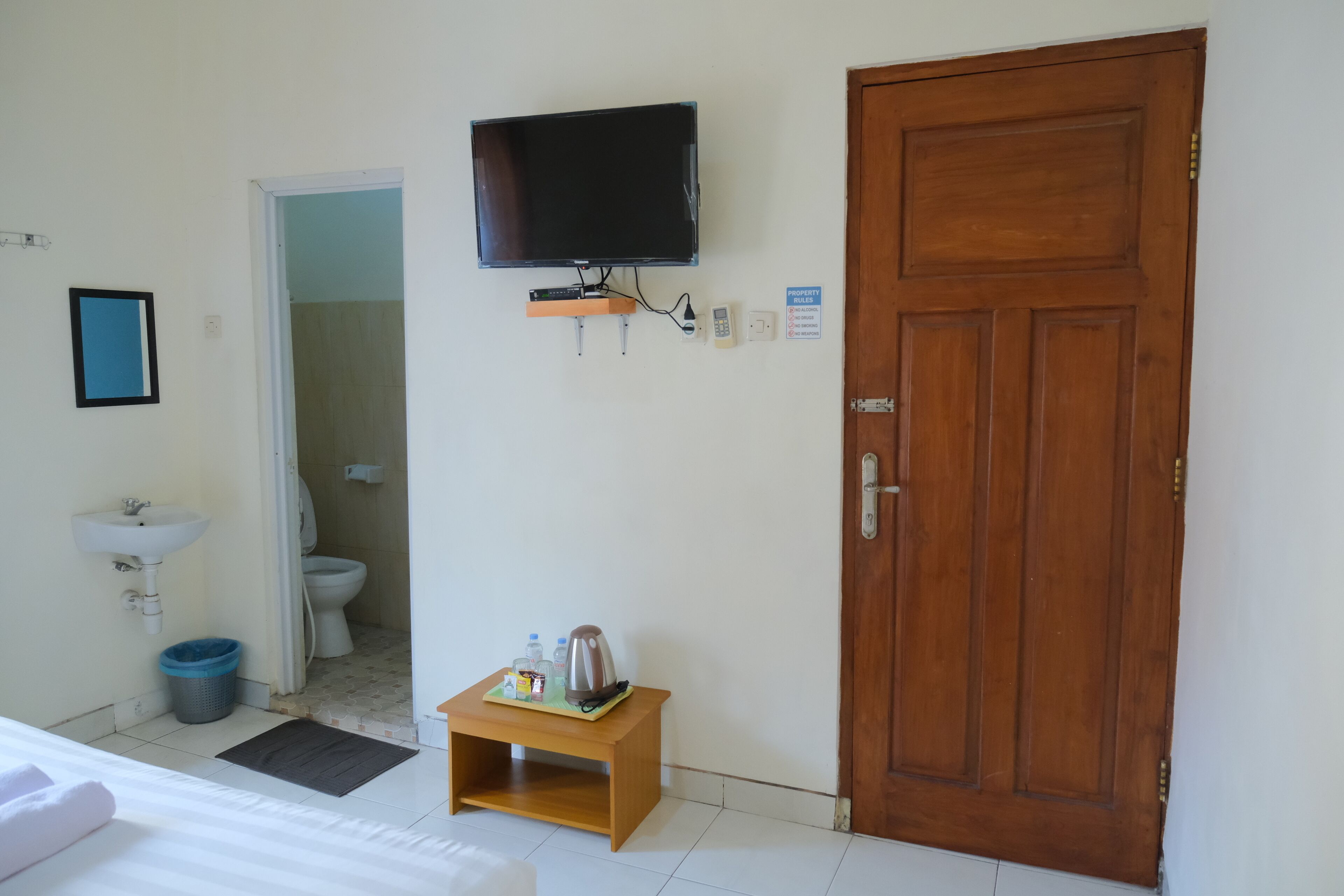 A hotel room with a wooden door, a television mounted on the wall, a small wooden table with a kettle and bottles, a sink, and a blue trash bin.