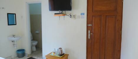 A hotel room with a wooden door, a television mounted on the wall, a small wooden table with a kettle and bottles, a sink, and a blue trash bin.