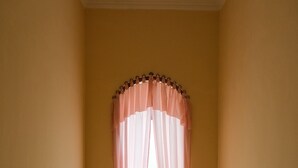Una habitación con una ventana cubierta por una cortina rosa, un pequeño estante con objetos y un suelo de baldosas.