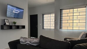 Una sala de estar moderna con un sofá gris oscuro, un televisor de pantalla plana montado en la pared y una ventana con persianas.