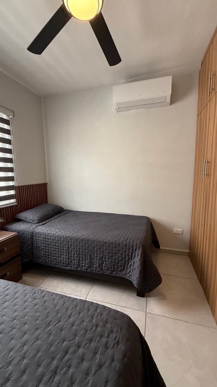 Un dormitorio con dos camas, ventilador de techo, equipo de aire acondicionado, armario de madera y ventana con persianas.