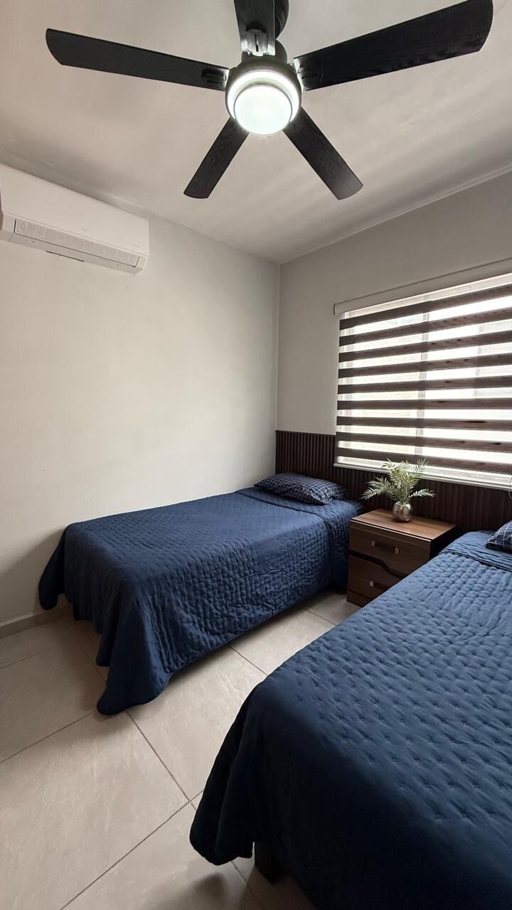 A bedroom with two beds, a ceiling fan, a window with blinds, and a bedside table with a plant.