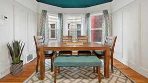 A dining area with a wooden table, four chairs, and a bench. There are patterned curtains on the windows, a potted plant, and a thermostat on the wall. - Newly renovated 2-bedroom apartment in quiet Historic Detroit neighborhood (Detroit)
