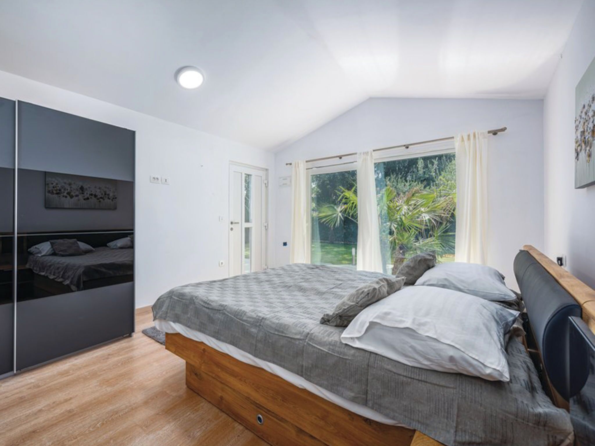 A bedroom with a bed, a wardrobe, a window with curtains, and a view of greenery outside.