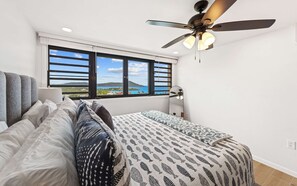 A bedroom with a bed, pillows, a ceiling fan, a nightstand, a lamp, and a window with a view of the sea. - 2 Pools, Dining | Walk to Beach | Sapphire Village (St. Thomas)