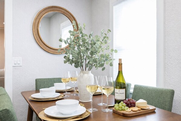 Een eettafel gedekt met witte borden, gouden glazen en een fles Napa Valley wijn, met een vaas met groene bladeren.
