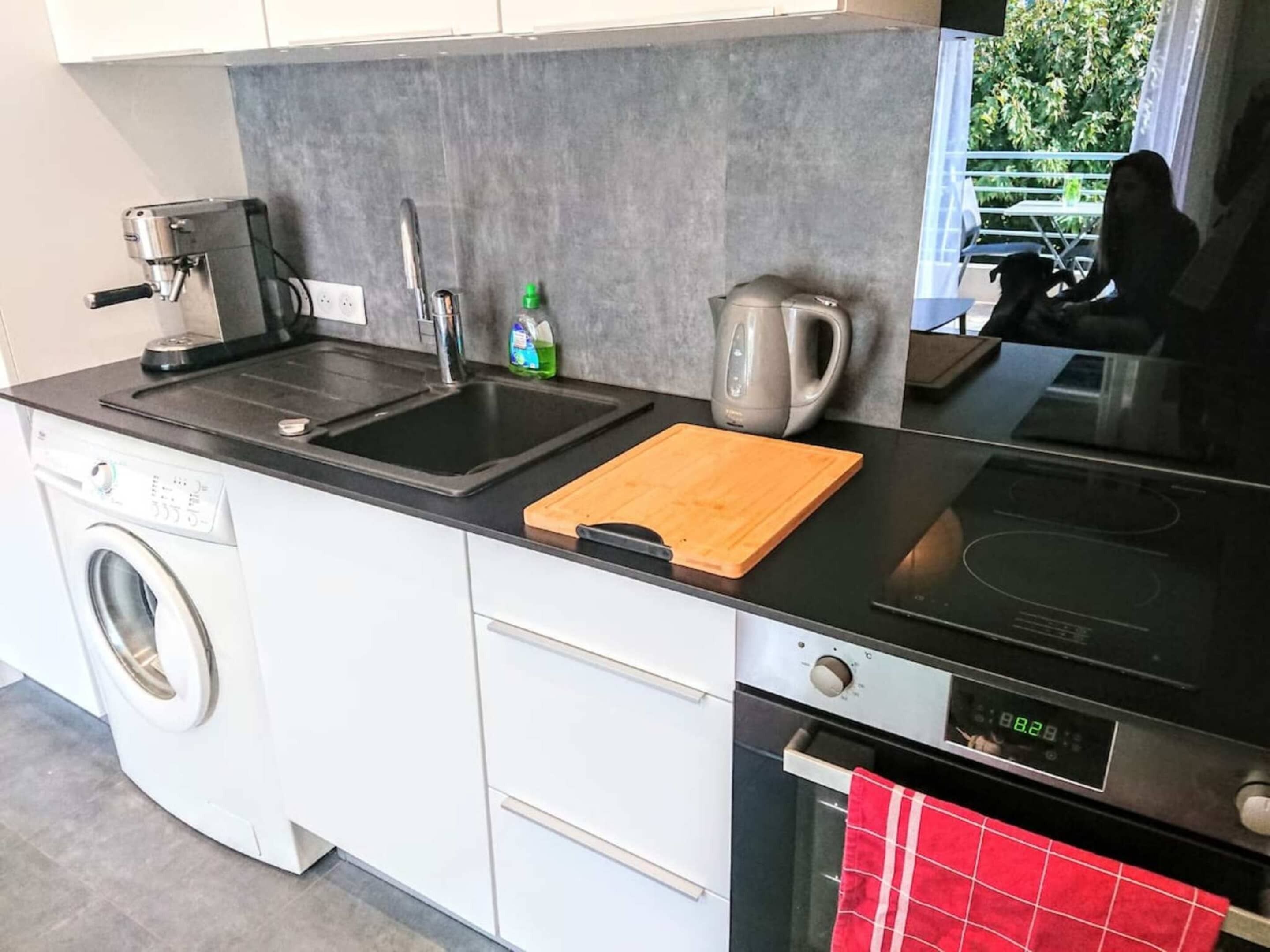A compact kitchen with a washing machine, a coffee maker, a kettle, and a red towel hanging on the oven door.