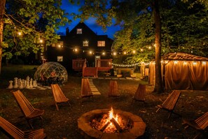 A nighttime outdoor setting with a fire pit, wooden chairs, string lights, and a house in the background. - Wellness Escape I Sauna, Hot Tub, Theater & Game Room (East Stroudsburg)