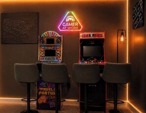 A gaming area with arcade machines, a neon sign reading 'GAMER AT WORK,' and a 'NBA JAM' game.