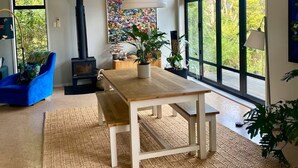 A modern living room with a wooden dining table, a blue armchair, a large window with a view of greenery, a fireplace, and a ceiling fan. - Omiha Exclusive Retreat (Waiheke Island)
