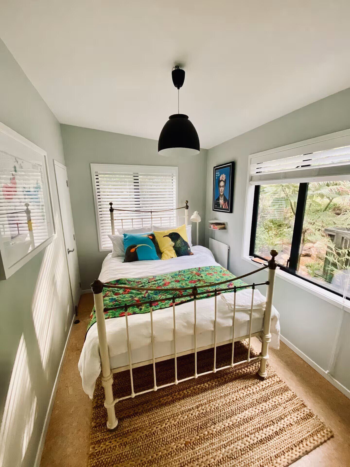 A bedroom with a bed, a window with blinds, a hanging light fixture, and a view of greenery outside.