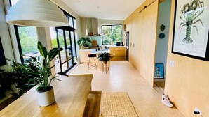 A modern kitchen with a wooden island, large windows, and a dining area with a rug. - Omiha Exclusive Retreat (Waiheke Island)