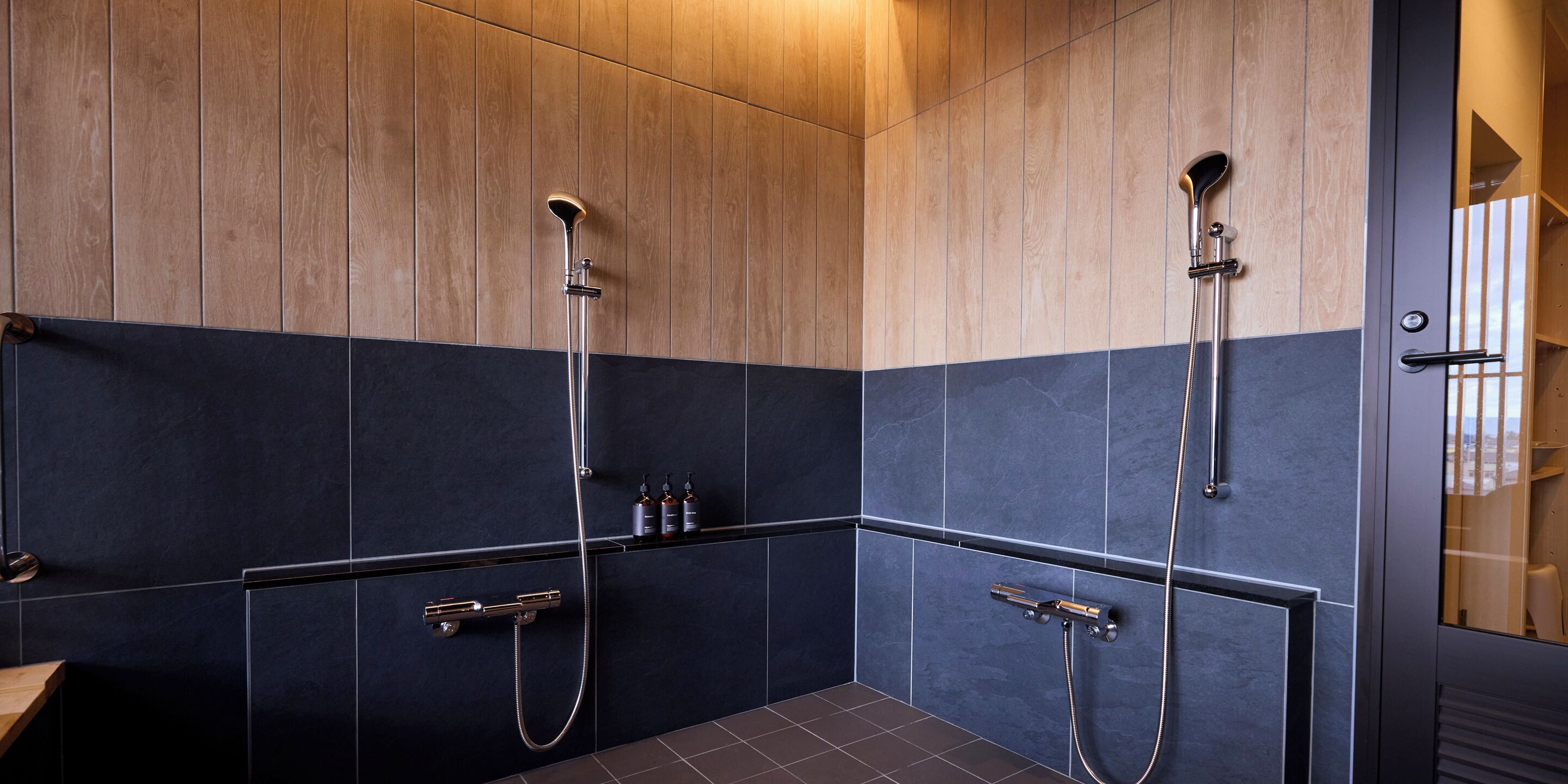 A modern bathroom with two showerheads, dark tiles, and wooden paneling.