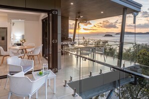 A modern outdoor dining area with a view of the sea at sunset, featuring a glass railing and contemporary furniture. - Hidden Cove 17 (Hamilton Island)
