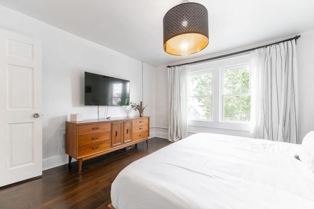 A bedroom with a large bed, a wooden dresser, a flat-screen TV, and a window with curtains.