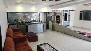 A hotel reception area with a reception desk, a seating area with sofas, a small table with a newspaper, and a staircase leading to the restaurant. - Hotel Palika (Hassan)