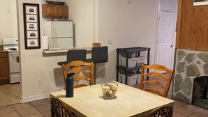 A dining area with a table and chairs, a ceiling fan, a kitchen in the background, and a stone fireplace.