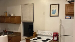 A kitchen with a stove, oven, and refrigerator. There is a window with blinds, a sink, and wooden cabinets.