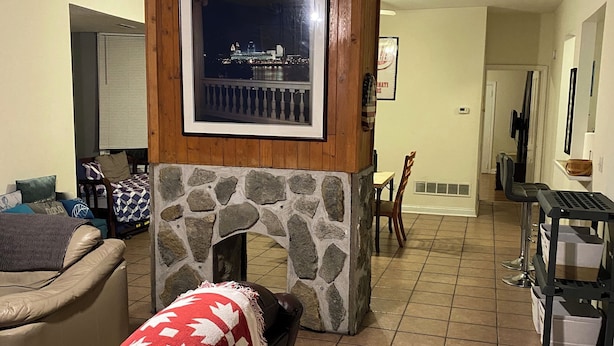 A living room with a stone fireplace, a framed picture above it, a couch with a red and white patterned throw, and a tiled floor.