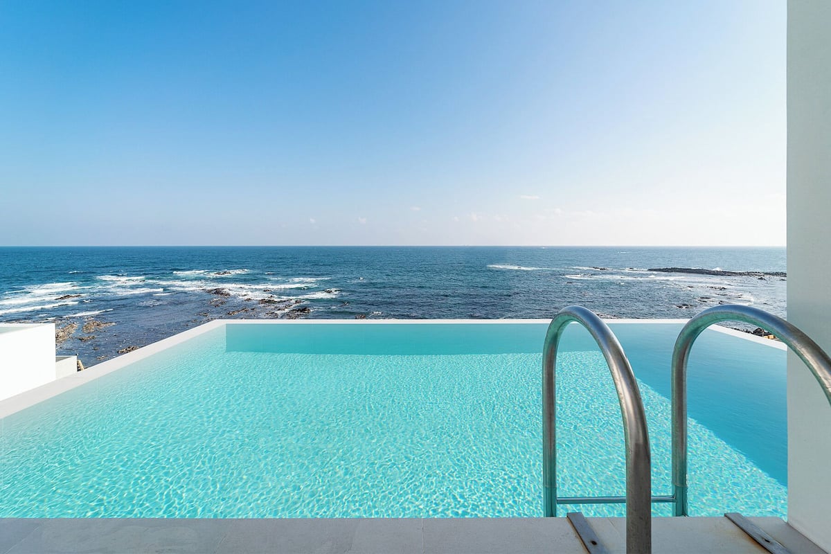 A clear blue infinity pool with metal railings, overlooking a rocky coastline and the ocean.