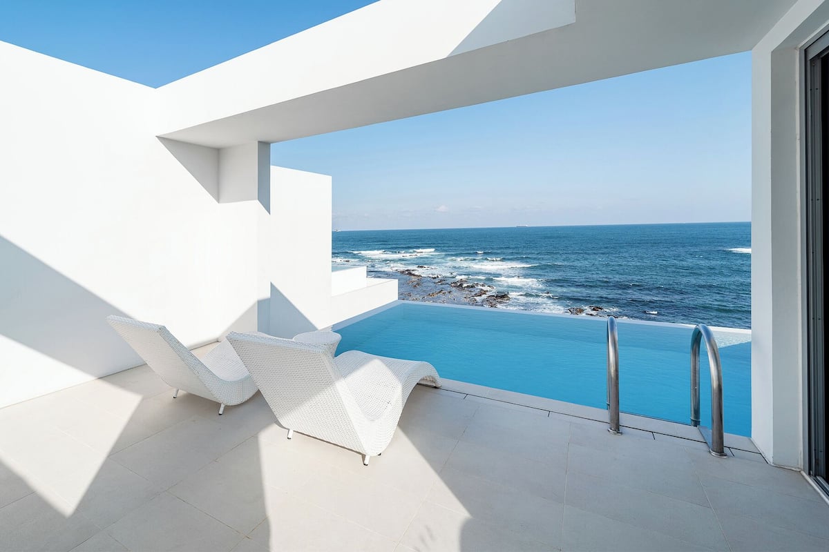 A modern outdoor pool area with white wicker furniture and a view of the ocean.