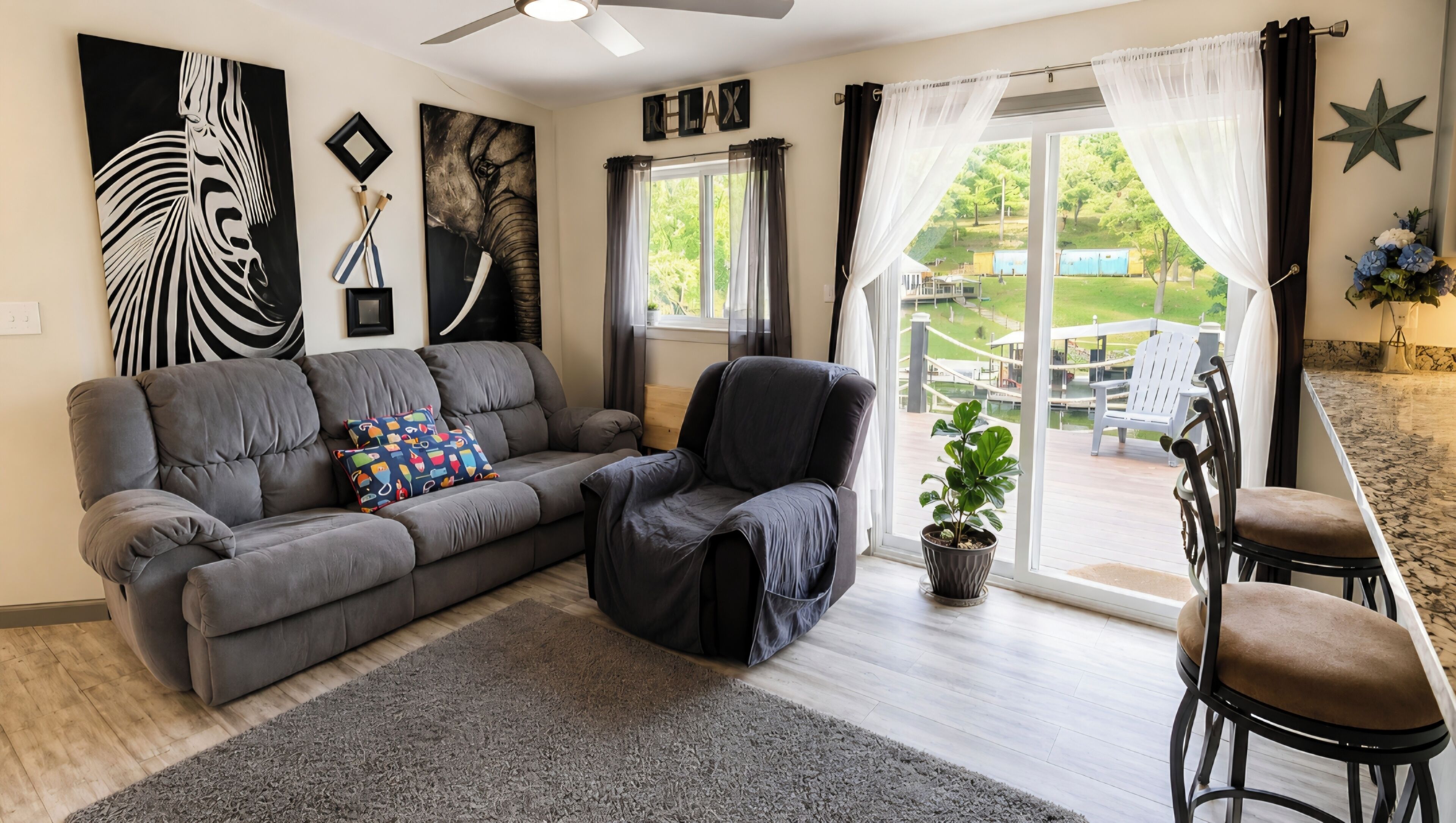 A living room with a grey sofa, a black armchair, a patterned throw pillow, a potted plant, and a view of a deck with outdoor furniture.