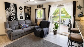 A living room with a grey sofa, a black armchair, a patterned throw pillow, a potted plant, and a view of a deck with outdoor furniture.