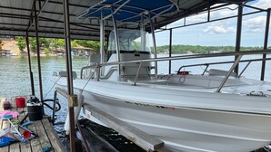 A boat docked at a pier with a covered area overhead, a wooden deck with various boating equipment, and a view of a calm body of water with greenery in the background.
