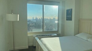 A hotel room with a bed, a floor lamp, a radiator, a window with a city view, and a framed picture on the wall.