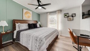 A bedroom with a bed, bedside tables, a desk, a chair, a ceiling fan, and a window with curtains. - Newly Remodeled Mobile Home The Green Showcase! (Mineral Wells)