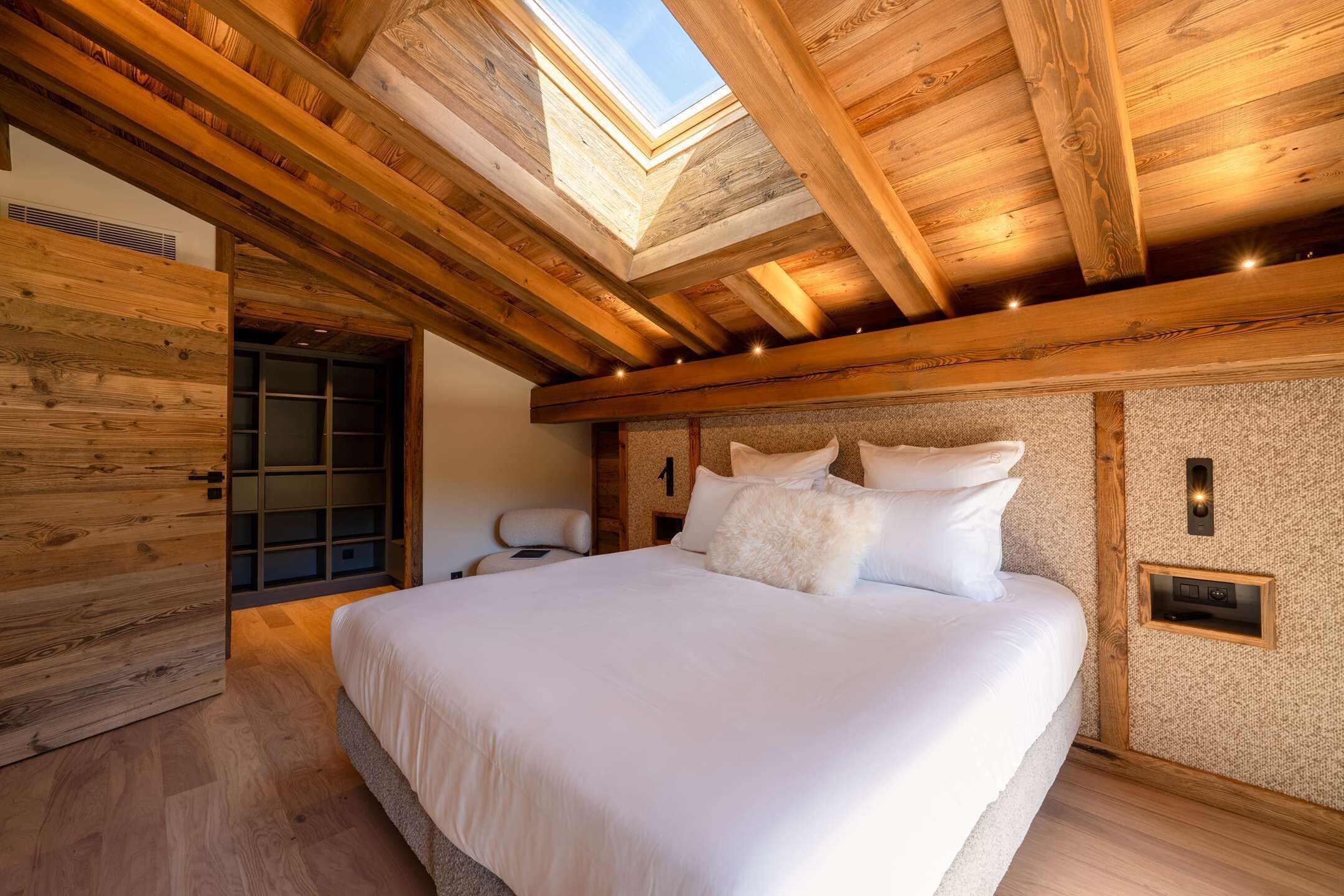 A bedroom with a skylight, wooden beams, a bed with white bedding, and a small bedside table.