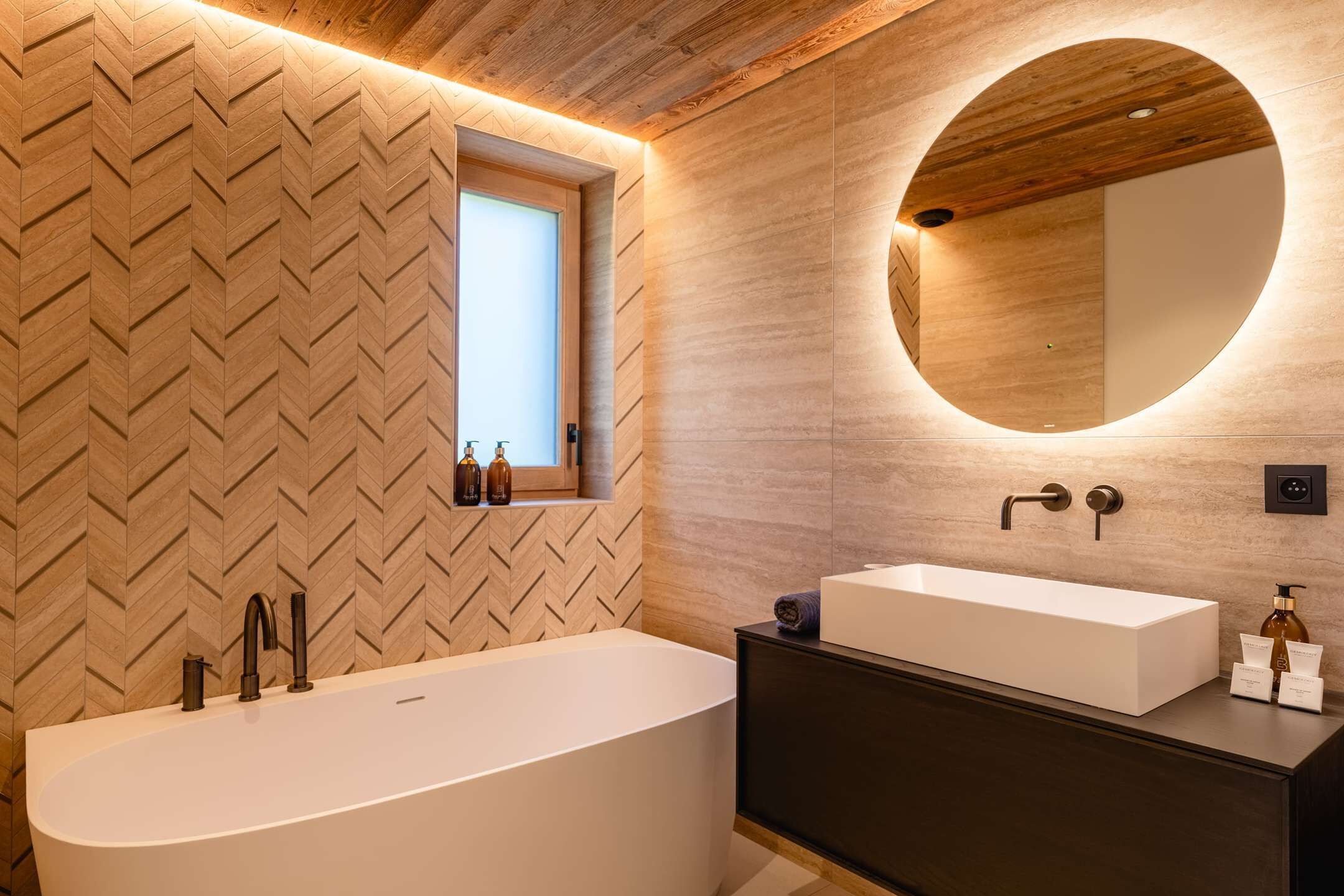 Een moderne badkamer met een badkuip op poten, een rond spiegeltje, een donker badmeubel en een witte wastafel. De wand is bekleed met hout in een herringbone-patroon.