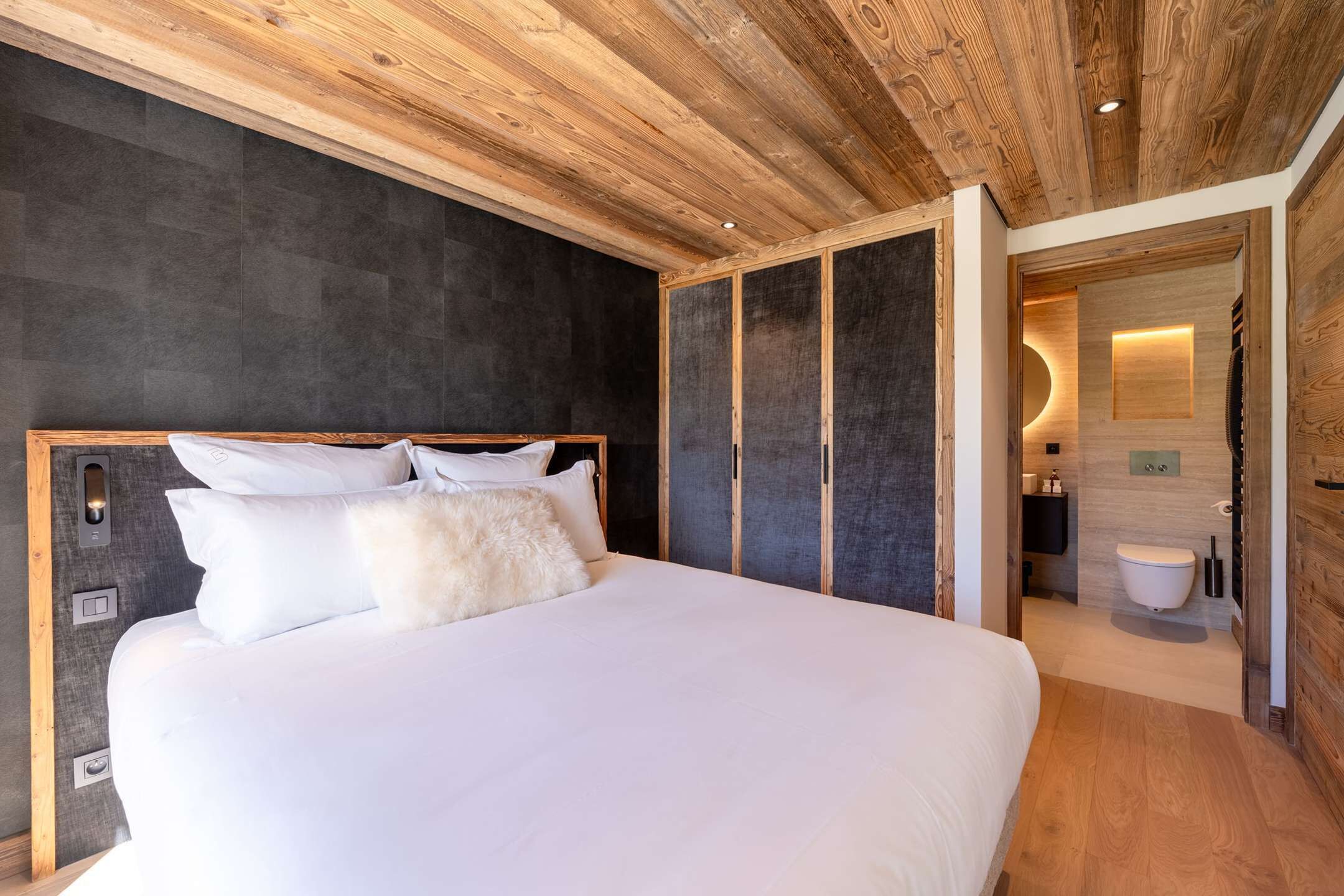 A modern bedroom with a large bed, dark walls, and wooden ceiling.