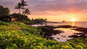 Paisaje costero con una casa, palmeras y un barco al atardecer.