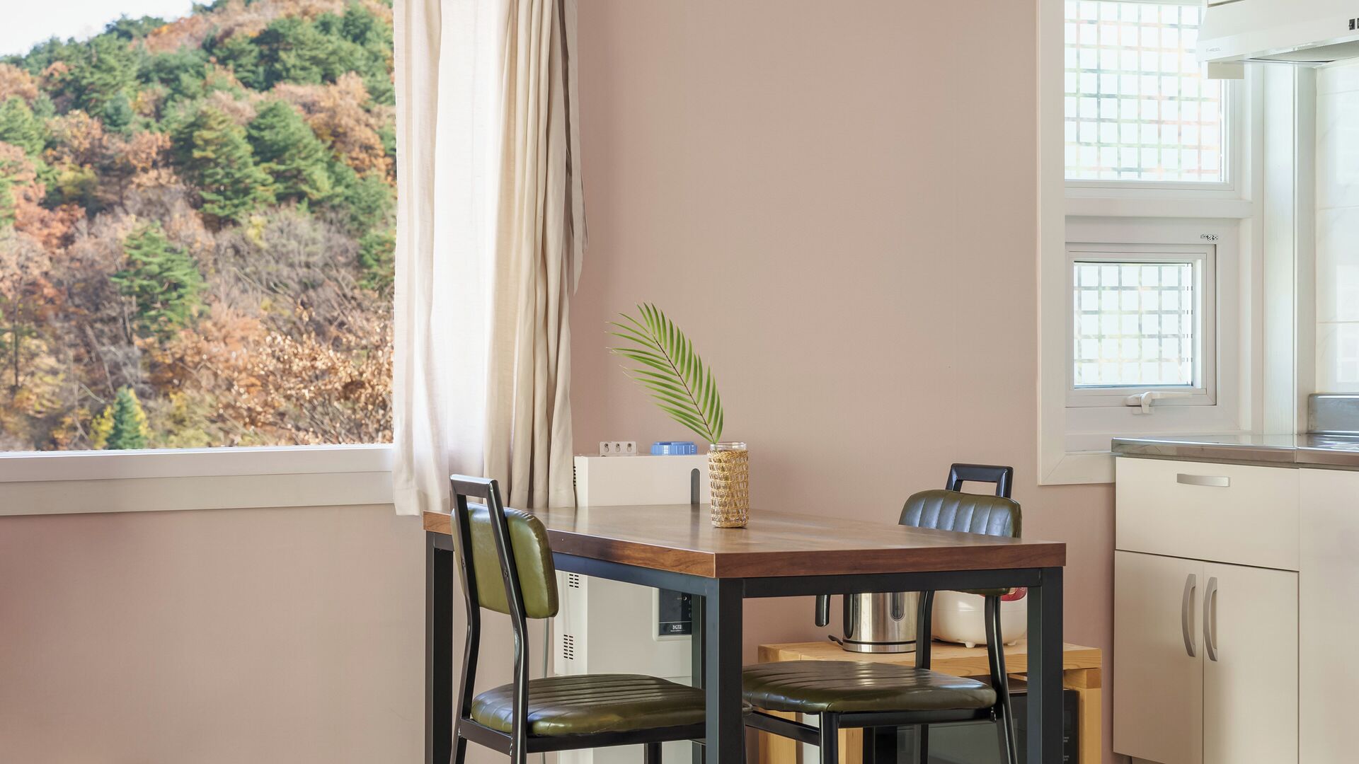 Een keuken met een eettafel, stoelen, een plant en uitzicht op een bos.